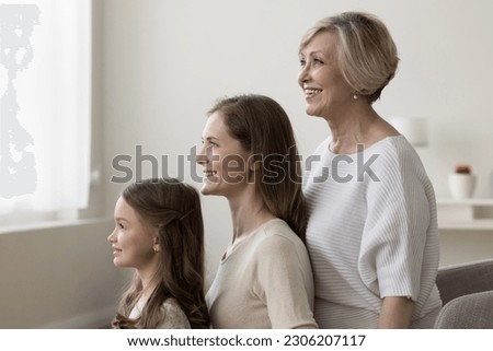 Young beautiful woman, her elderly pensioner mother and adorable preschooler daughter standing in row in living room. Portrait of multi-generational women family. Heredity, offspring, next generation