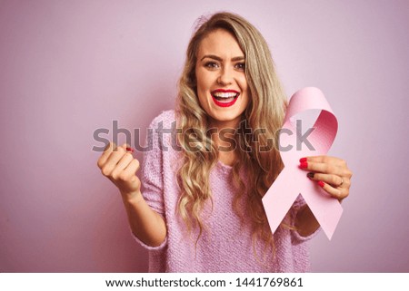 Young beautiful woman holding cancer ribbon over pink isolated background screaming proud and celebrating victory and success very excited, cheering emotion