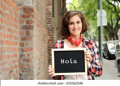 Young beautiful woman holding chalkboard with text "Hola". Outdoors.