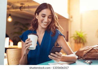 Young beautiful woman holding coffee paper cup and looking at smartphone while sitting at cafeteria. Happy university student girl using mobile phone. Businesswoman drinking coffee and smiling.