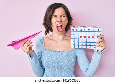 Young Beautiful Woman Holding Heart Calendar And Paper Airplane Angry And Mad Screaming Frustrated And Furious, Shouting With Anger. Rage And Aggressive Concept. 