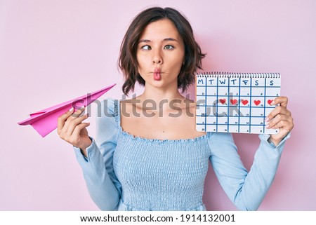 Young beautiful woman holding heart calendar and paper airplane making fish face with mouth and squinting eyes, crazy and comical. 