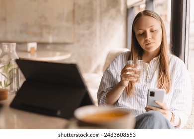 Young beautiful woman holding mobile and looking at smartphone while sitting at cafeteria. Calm girl using mobile phone and look flirting. Woman drinking water and smiling, sitting in cafe. - Powered by Shutterstock