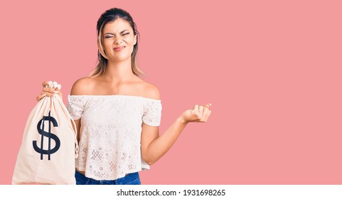 Young Beautiful Woman Holding Money Bag With Dollar Symbol Annoyed And Frustrated Shouting With Anger, Yelling Crazy With Anger And Hand Raised 