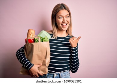 Young Beautiful Woman Holding Paper Bag With Purchase Over Isolated Pink Background Pointing And Showing With Thumb Up To The Side With Happy Face Smiling