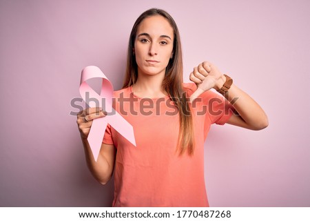 Young beautiful woman holding pink cancer ribbon standing over isolated pink background with angry face, negative sign showing dislike with thumbs down, rejection concept
