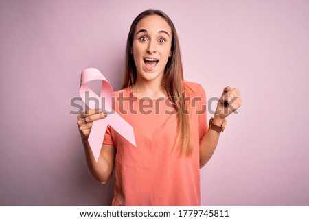 Young beautiful woman holding pink cancer ribbon standing over isolated pink background screaming proud and celebrating victory and success very excited, cheering emotion