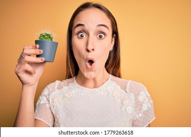 Young beautiful woman holding small cactus plant pot over isolated yellow background scared in shock with a surprise face, afraid and excited with fear expression