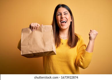 Young beautiful woman holding take away paper bag from delivery over yellow background screaming proud and celebrating victory and success very excited, cheering emotion - Shutterstock ID 1673922874