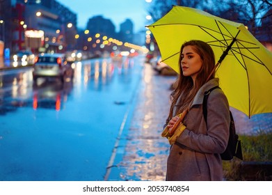 Young beautiful woman holding a yellow umbrella while waiting for a cab on a rainy night near the road