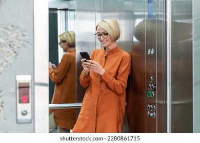 Young beautiful woman inside office building at workplace working, businesswoman using phone standing in elevator, smiling and happy female worker starts new successful day.
