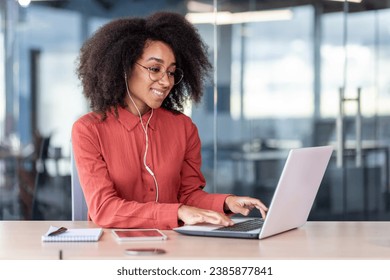 Young beautiful woman inside the office works with a laptop, a businesswoman in headphones listens music, podcasts, audio books and training course. Worker smiling , with curly hair and red shirt. - Powered by Shutterstock