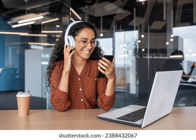 Young beautiful woman inside office with phone in hands and headphones listening to music and watching online video using application on smartphone, businesswoman relaxing at workplace with laptop. - Powered by Shutterstock