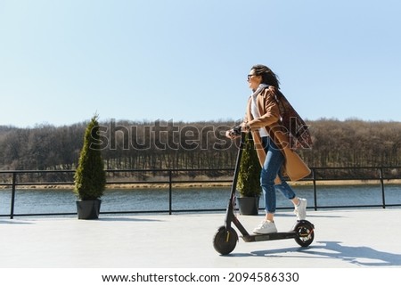 Young beautiful woman in a jacket smiles and rides an electric scooter to work along office buildings