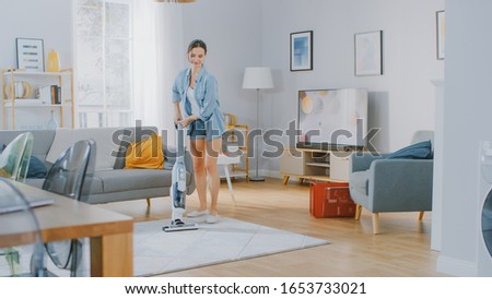 Young Beautiful Woman in Jeans Shirt and Shorts is Vacuum Cleaning a Carpet in a Bright Cozy Room at Home. She Uses a Modern Cordless Vacuum. She's Happy and Cheerful.