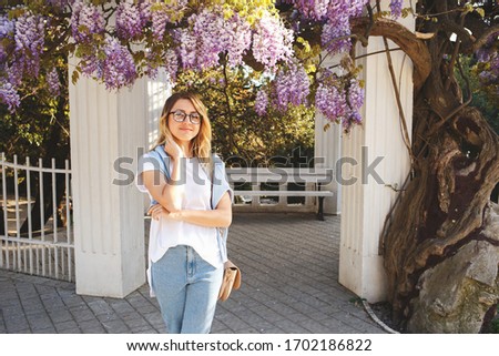 Young beautiful woman in jeans, white t-shirt and glasses stands near a flowering Wisteria tree. Spring portrait.