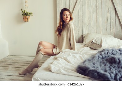 young beautiful woman in knitted cardigan and warm socks wake up in the morning in cozy scandinavian bedroom and sitting on bed with white bedlinen. Casual lifestyle in modern interior