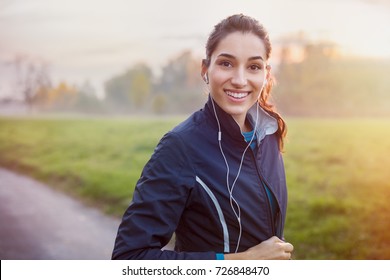 Young Beautiful Woman Listening Music At Park While Running. Portrait Of Smiling Sporty Girl With Earphone Looking At Camera At Park In Autumn. Woman Athlete Looking At Camera During Winter Sunset.