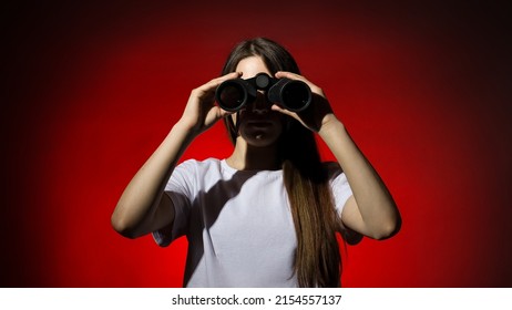 young beautiful woman with long hair in a white t-shirt looks through binoculars on a red background