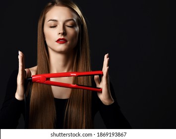Young beautiful woman with long silky straight hair in black clothes with eyes closed holding red hair straightener in hands over dark background. Haircare, beauty, wellness, styling concept