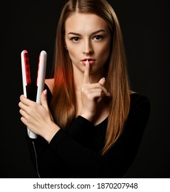 Young beautiful woman with long silky straight hair in black bodysuit holds hair straightener in hand and finger at lips gesturing silence sign over dark background. Haircare, beauty, wellness concept