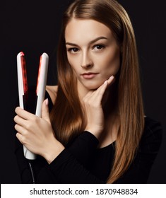 Young beautiful woman with long silky well-groomed straight hair in black bodysuit holding hand at her chin demonstrates hair straightener over dark background. Haircare, beauty, wellness concept