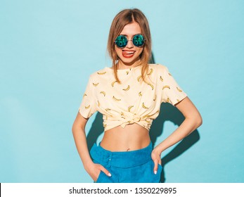 Young beautiful woman looking at camera. Trendy girl in casual summer yellow T-shirt clothes showing her tongue in round sunglasses. Positive female shows facial emotions. Funny model isolated on blue