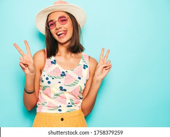 Young beautiful woman looking at camera in hat.Trendy girl in casual summer white T-shirt and yellow skirt in round sunglasses. Positive female shows facial emotions. Shows peace sign