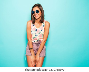 Young beautiful woman looking at camera.Trendy girl in casual summer white T-shirt and jeans shorts in round sunglasses. Positive female shows facial emotions. Funny model isolated on blue