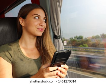 Young Beautiful Woman Looking Through The Bus Window. Happy Bus Passenger Traveling Sitting In A Seat And Looking Through The Window With Smart Phone In Her Hand.