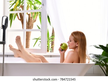 Young Beautiful Woman Lying In Bathtub And Taking Bath Near Open Bathroom Window Eating Green Apple 