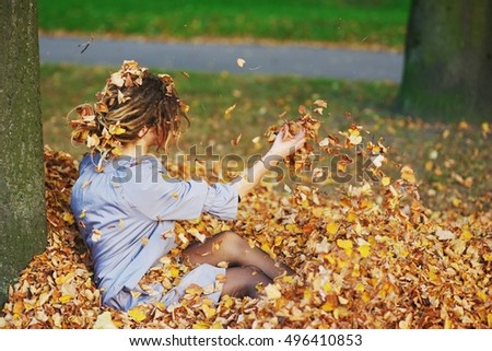 Young beautiful woman lying near a tree on a pile of fallen autumn leaves and throws them into the air.