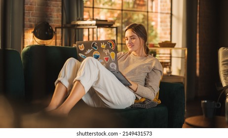 Young Beautiful Woman Lying on Couch, Using Laptop Computer in Stylish Loft Apartment on a Sunny Day. Creative Female Smiling, Checking Social Media, Typing Message. Urban City View from Big Window.