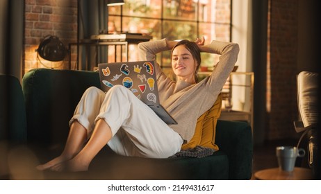 Young Beautiful Woman Lying on Couch, Using Laptop Computer in Stylish Loft Apartment on a Sunny Day. Creative Female Smiling, Checking Social Media, Typing Message. Urban City View from Big Window.