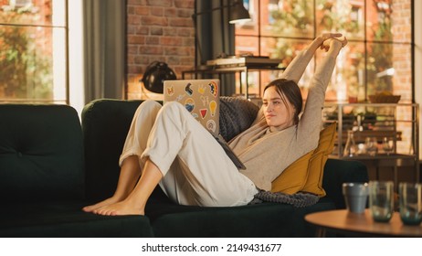Young Beautiful Woman Lying on Couch, Using Laptop Computer in Stylish Loft Apartment on a Sunny Day. Creative Female Smiling, Checking Social Media, Stretching. Urban City View from Big Window.