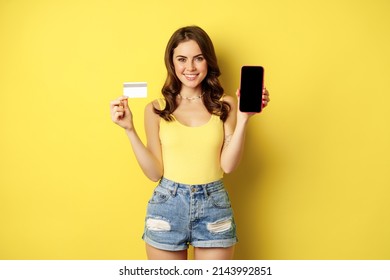 Young beautiful woman model showing smartphone empty phone screen and credit card, ready for summer, wearing tank top and shorts, standing over yellow background - Powered by Shutterstock