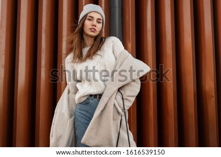 Young beautiful woman model in stylish youth warm outerwear posing near a modern wall on the street. Attractive girl in fashionable clothes is standing in the city. New spring collection. Style.