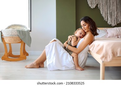 Young Beautiful Woman Mother Hugging Embracing Cute Baby Boy While Sitting Together On Floor In Bedroom Near Bassinet. Mom Cuddling Her Little Son Infant And Expressing Love And Care.