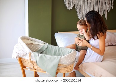 Young Beautiful Woman Mother With Sleepy Baby Boy Son Sitting On Bed Near Bassinet, Loving Mom Putting To Sleep Cute Infant Rubbing His Eyes. Motherhood Concept