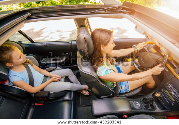Young beautiful woman with\
a nice smile driving a car.  Her cute daughter sitting on rear and\
enjoying.