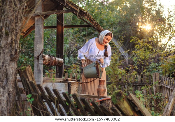 A\
young beautiful woman in old peasant clothes and a head scarf takes\
water from the well and carries it on a yoke in buckets, domestic\
female labor, rural life, historical\
reconstruction.