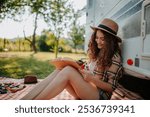 Young beautiful woman is on a camping trip in nature, sitting in front caravan and reading book.