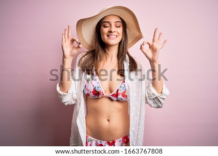 Young beautiful woman on vacation wearing bikini and summer hat over pink background relax and smiling with eyes closed doing meditation gesture with fingers. Yoga concept.