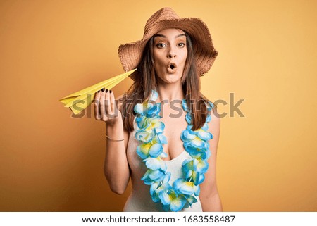 Young beautiful woman on vacation wearing bikini and hawaiian lei holding paper airplane scared in shock with a surprise face, afraid and excited with fear expression