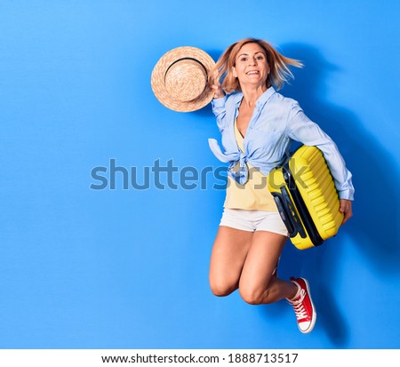 Young beautiful woman on vacation wearing summer clothes smiling happy. Jumping with smile on face holding cabin bag and summer hat over isolated background