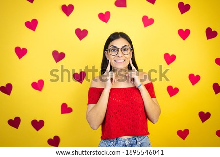 Young beautiful woman over yellow background with red hearts smiling confident showing and pointing with fingers teeth and mouth
