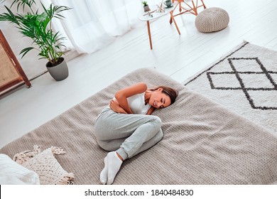 Young beautiful woman in painful expression holding her belly suffering menstrual period pain lying sad on home couch having tummy cramp in female health concept. Top view.