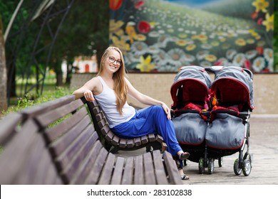 Young Beautiful Woman In A Park With A Double Jogging Stroller With Two Kids, Her Identical Twin Daughters