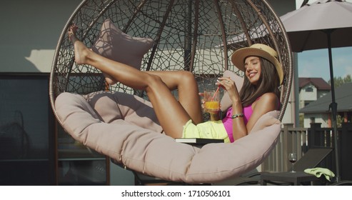 Young beautiful woman in pink bikini on hammock on terrace at luxury villa. Pretty girl in hat with notebook relaxing on pool edge. Female model drinking orange juice