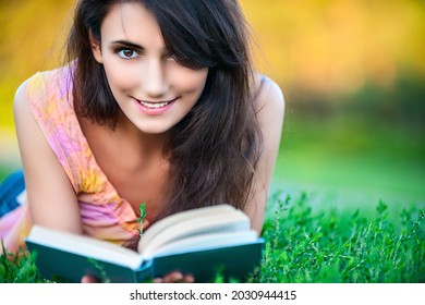 Junge schöne Frau in einer rosafarbenen Bluse und langes dunkles Haar liegt auf grünem Gras in einem Stadtpark und liest ein Buch.